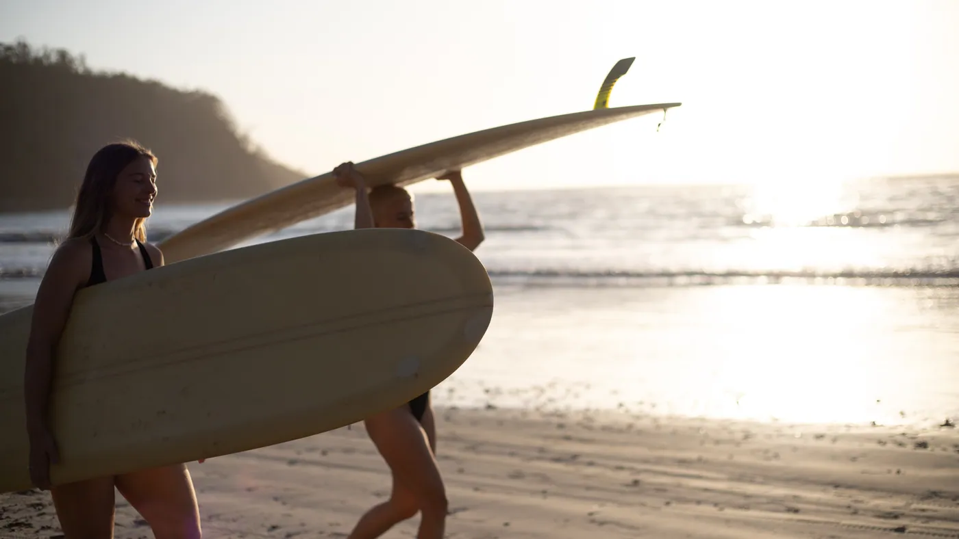 Two surfers carrying a board along the shoreline at golden hour