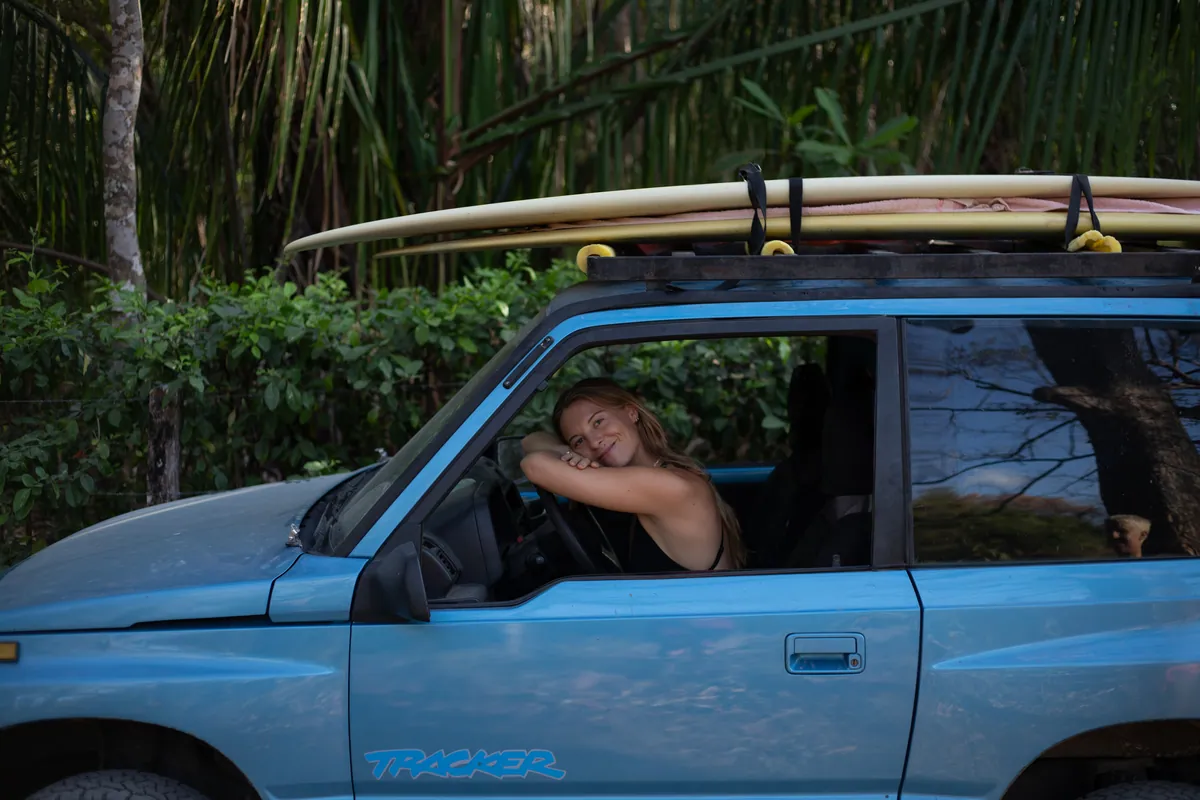 Charlotte in a blue truck loaded with surfboards, parked under palms